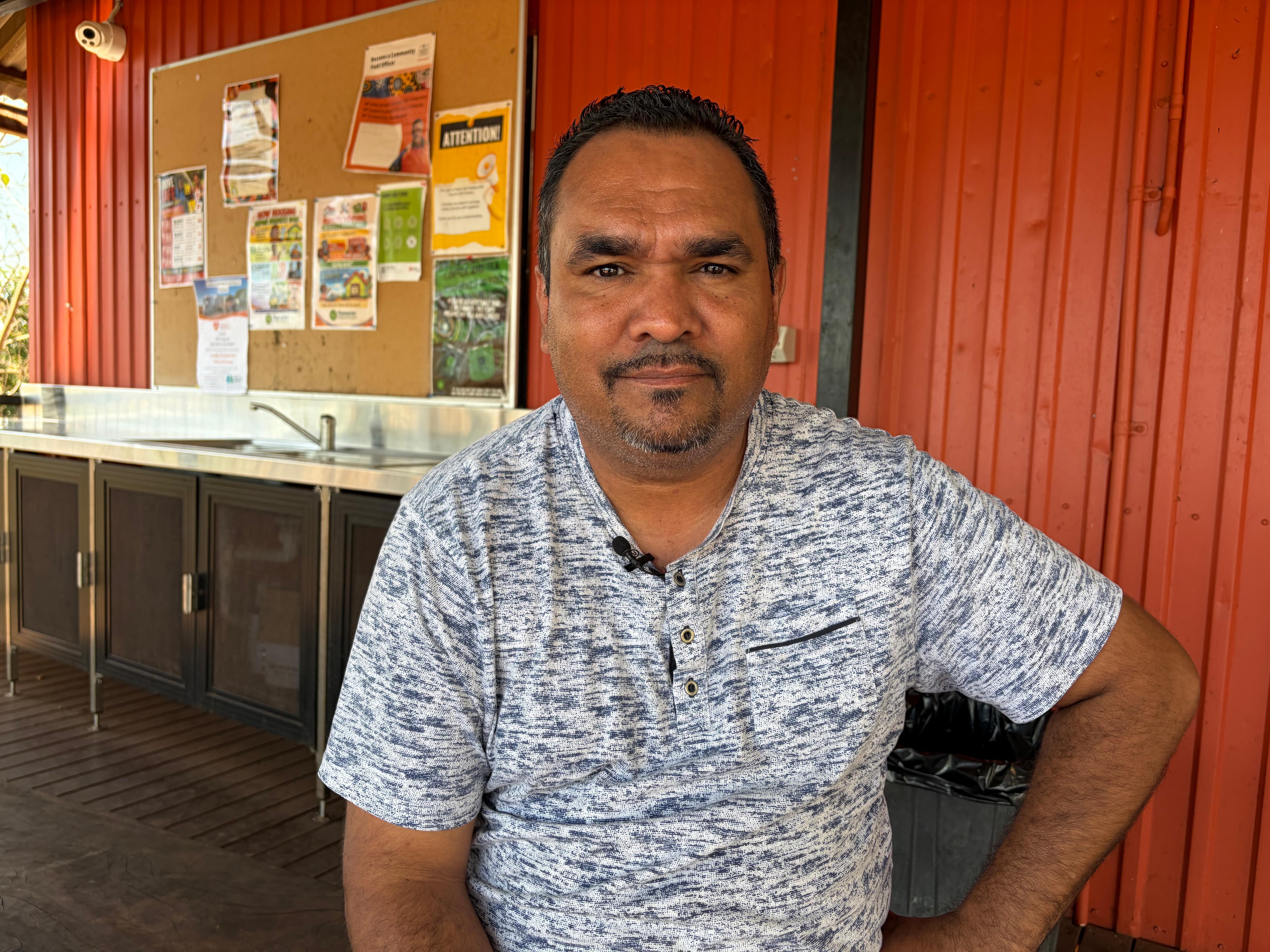 Mark Tunmuck Smith in a blue and white shirt with one hand on his hip looks straight ahead with a red wall in the background