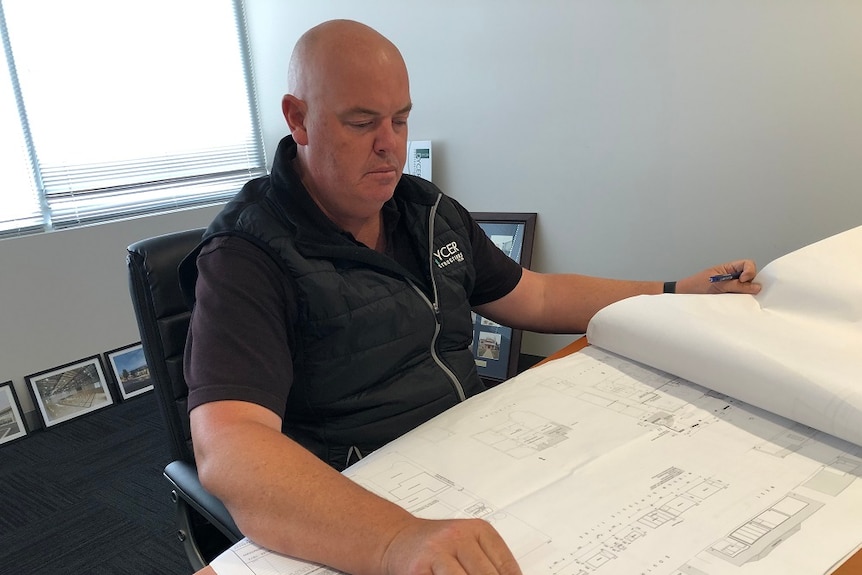 A bald man in a vest sits in a chair in an office with building plans and a ruler in front of him