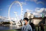 A man wears a mask and a denim jacket, the London Eye in the background.