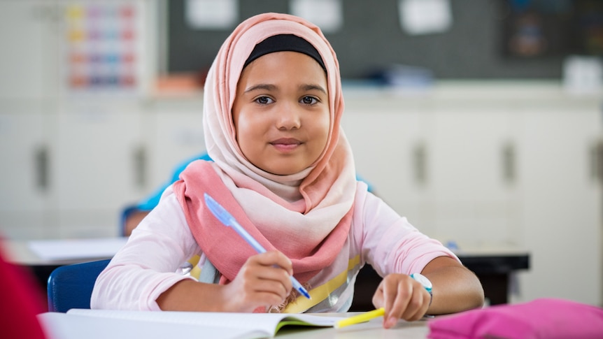 A young girl wearing a head covering sits in a classroom and smiles.