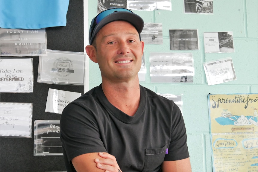 A man wearing a black shirt  and a black hat with blue sunglasses on top smiling in a classroom in front of a blue wall