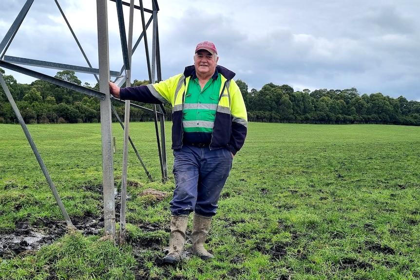 A man in gumboots stands in a wet paddock next to a transmission line.