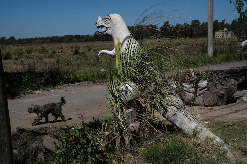 A dog walks on the road in front if a white dinosaur sculpture. 