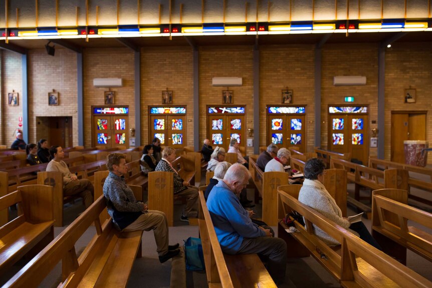 A small congregation sits in wooden pews in front of stained glass and stations of the cross.