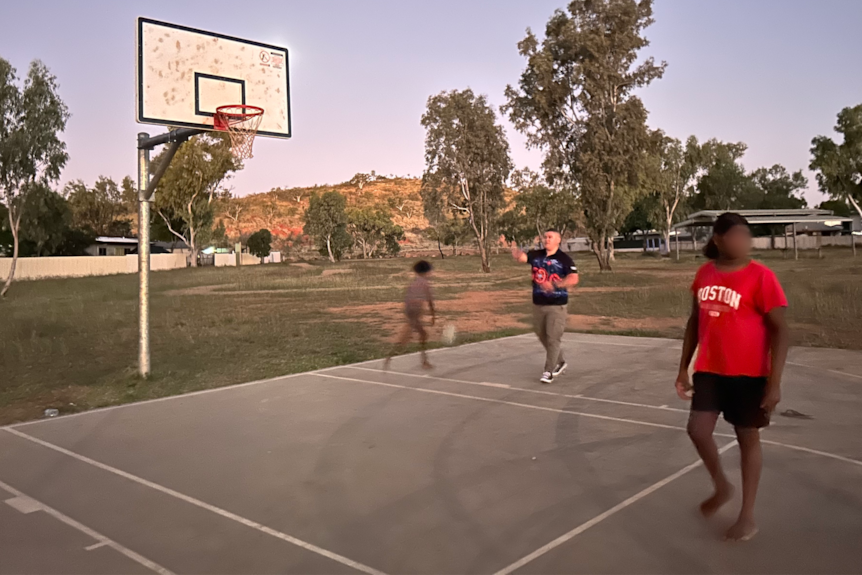 A man and two children play basketball at dusk. 