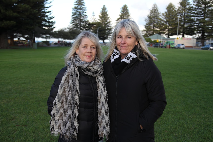Two blonde women in black puffer coats look somberly at the camera as they stand in a park