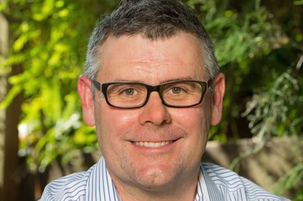 A man with a striped, collared short, short hair and glasses, smiling