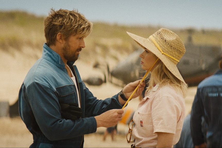 A man with sandy-coloured hair and a beard faces a woman wearing a large straw hat on a film set