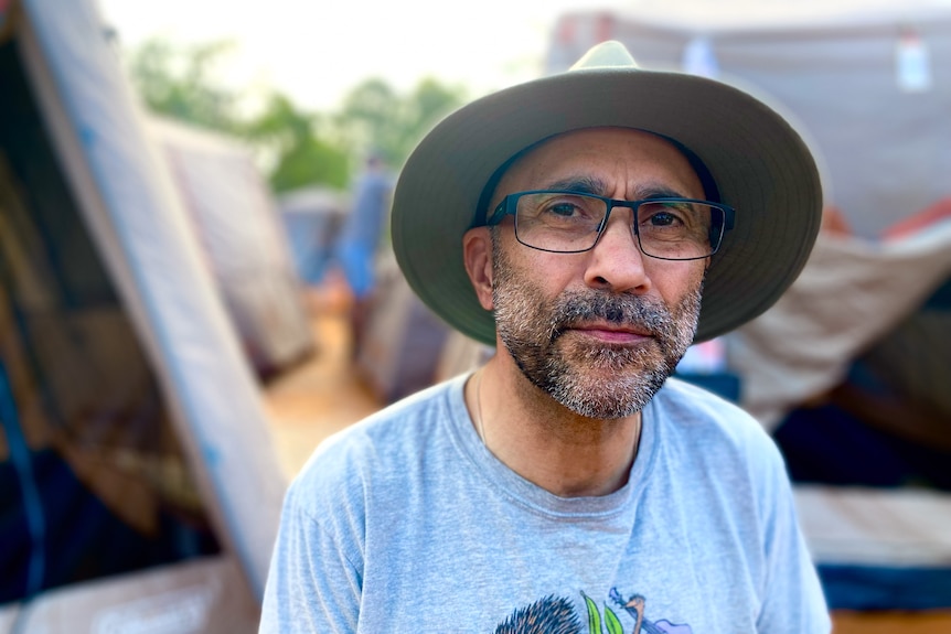 A bearded man wearing a hat and glasses with tents in the background.