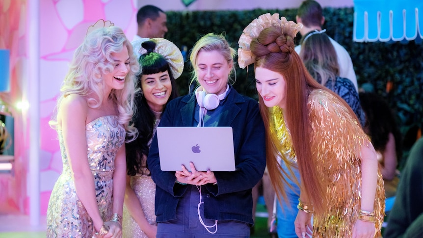 Actresses, dressed as Barbies, gather around Greta who is smiling and holding a laptop.