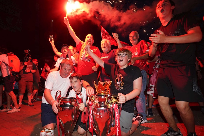 A group of fans dressed in red wave flares, cheer and shout standing next to replica trophies.