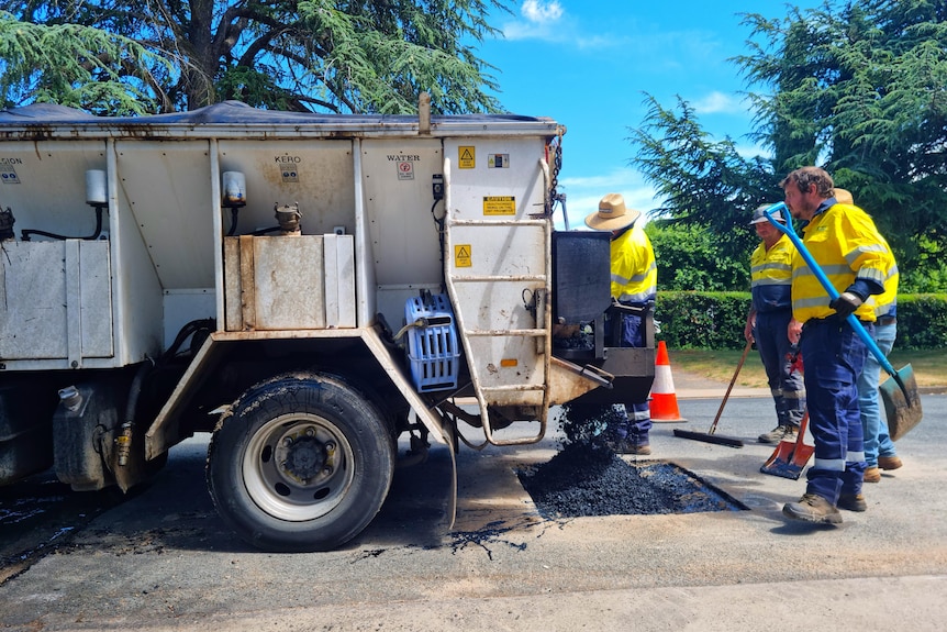 Three workers in high-vis clothing fill a pothole with bitumen behind a truck.