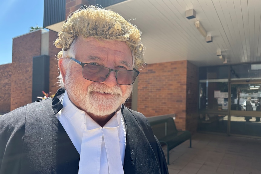 A smiling man in lawyer's garb, including wig, stands outside a court building.