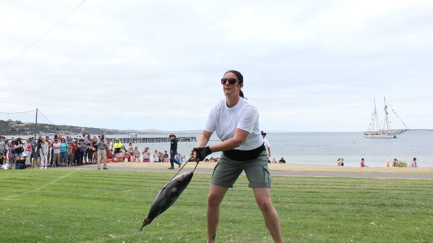 A woman in a white shirt and green shorts holding a frozen tuna attached to a rope which she is throwing onto the grass behind.