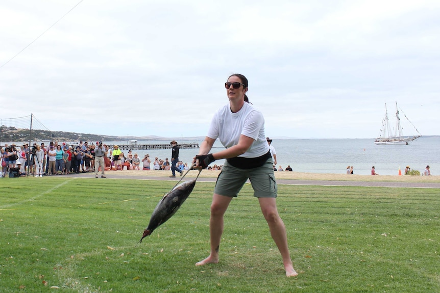 A woman in a white shirt and green shorts holding a frozen tuna attached to a rope which she is throwing onto the grass behind.
