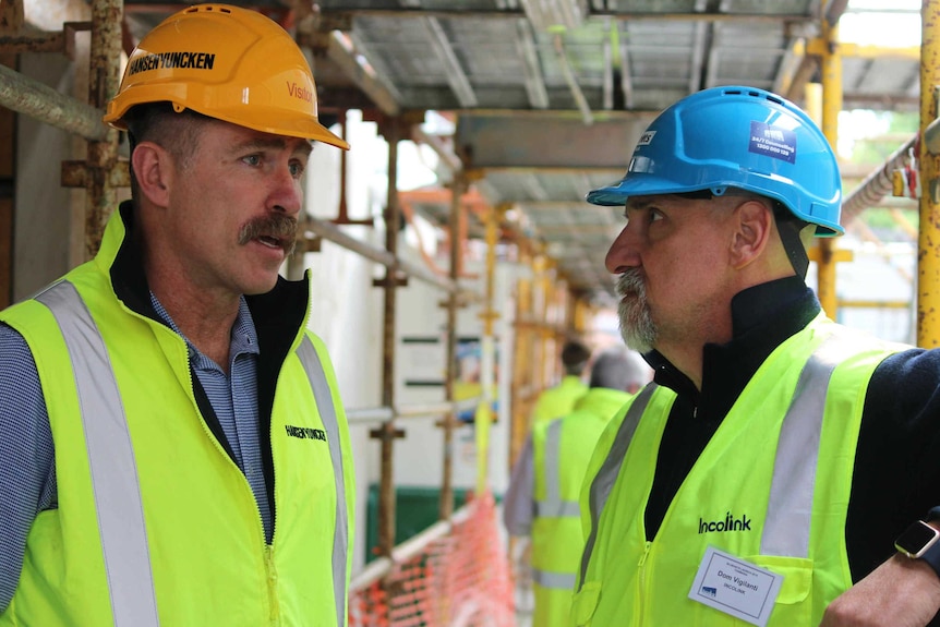 Two men in hard hats and hi-vis vests talk at a construction site
