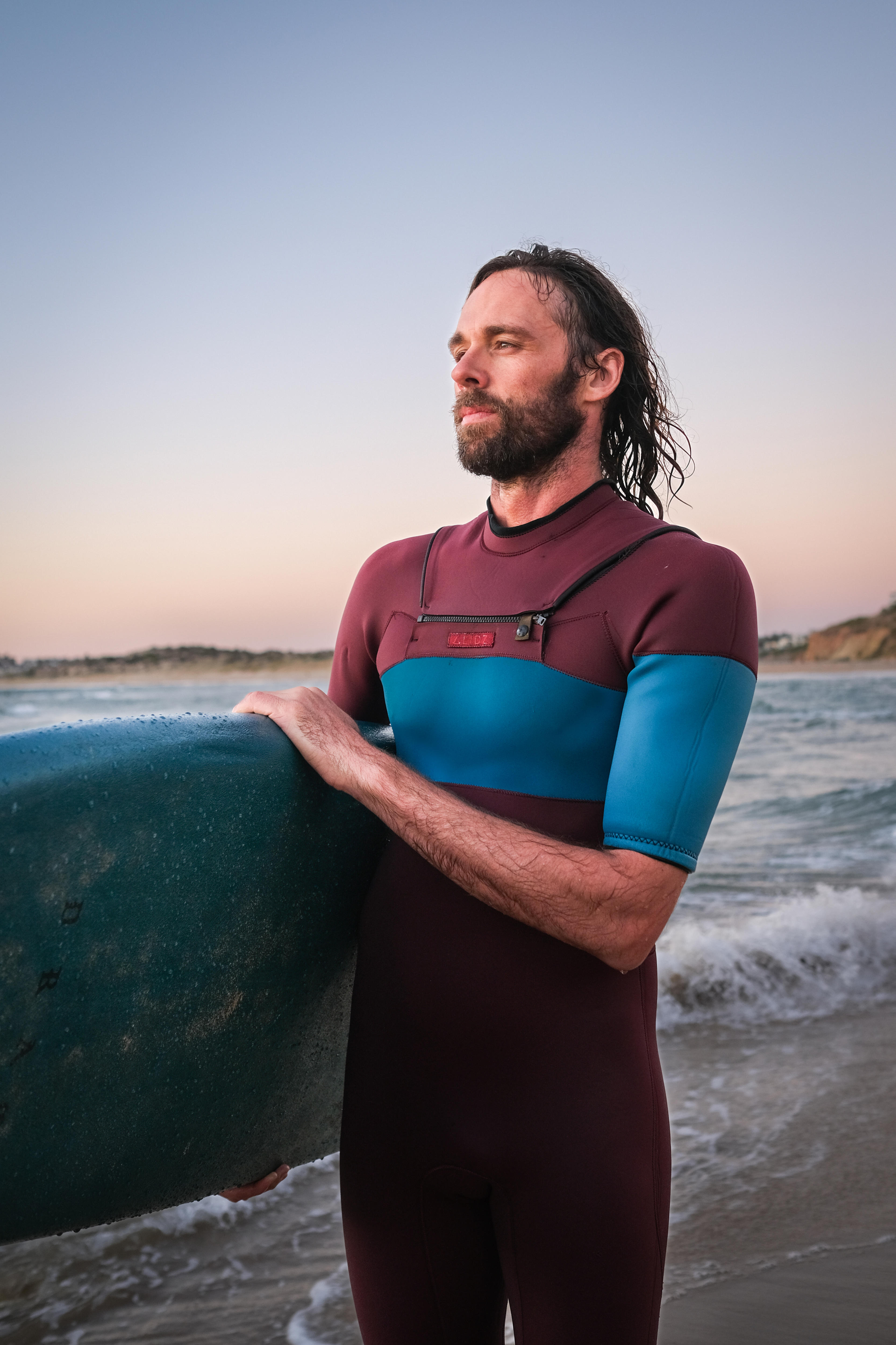 Henry Jock Walker stands next to the ocean after a surf, hair wet and wearing his wetsuit before sunset
