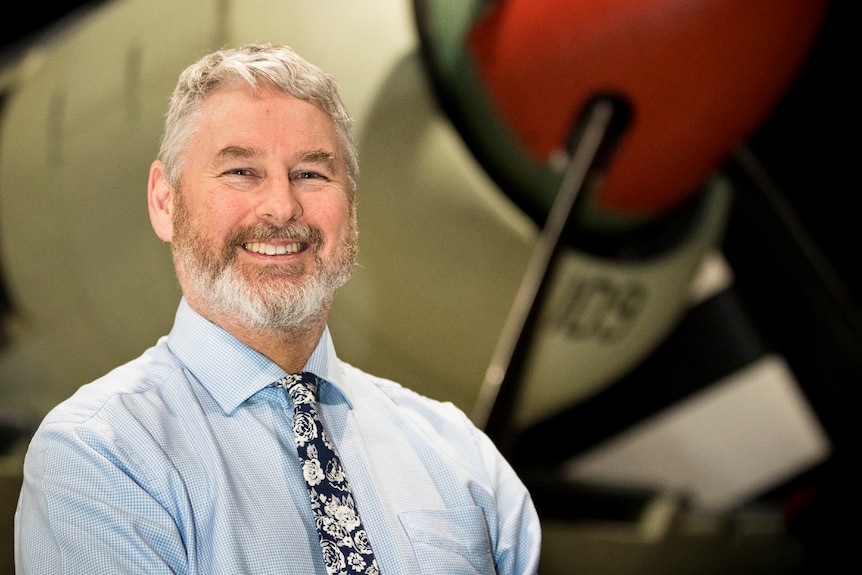 A smiling, silver-haired man with a neat beard stands in front of a World War II aircraft.