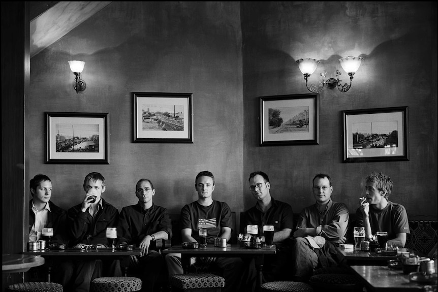 Seven men sit at a table in a pub with pints in front of them.