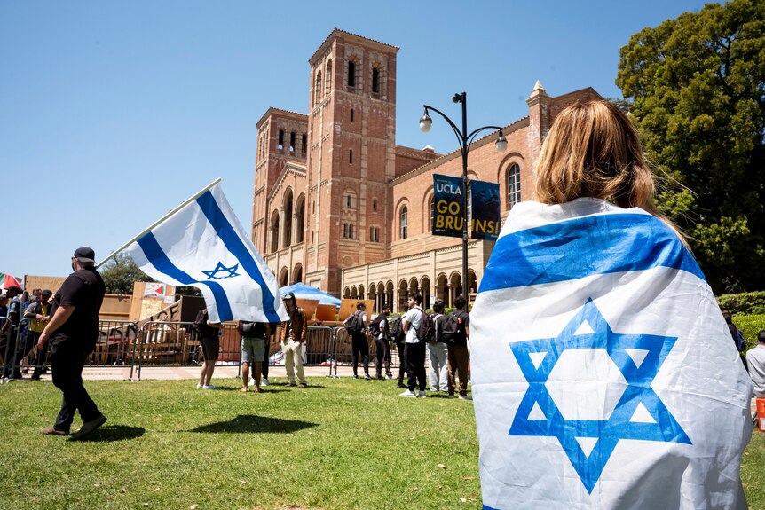 A woman wrapped in an Israeli flag stands with her back toward the camera in front of a university building
