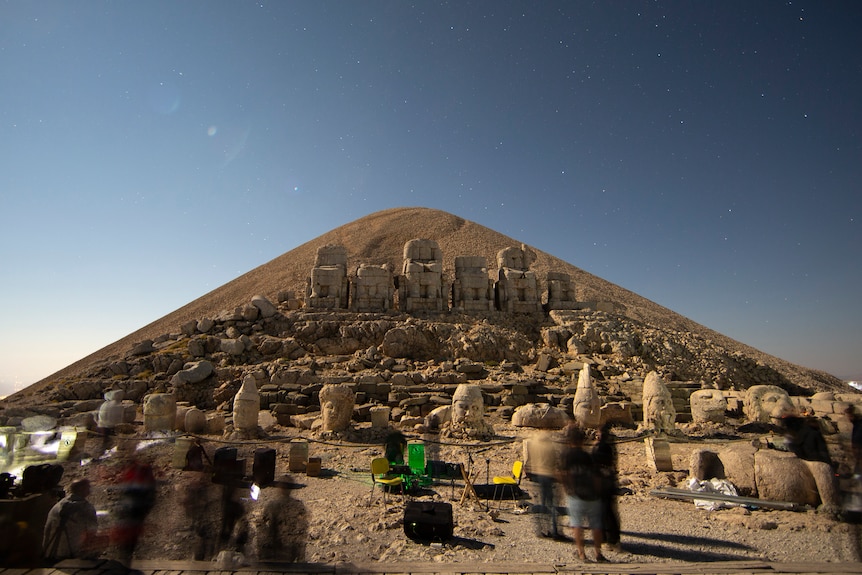 A long-exposure shot shows blurred people setting up camp on a mountaintop in front of ancient statues and a stunning night sky.