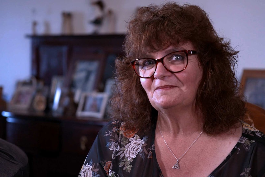 A woman in glasses looks at the camera with photo frames in the background.