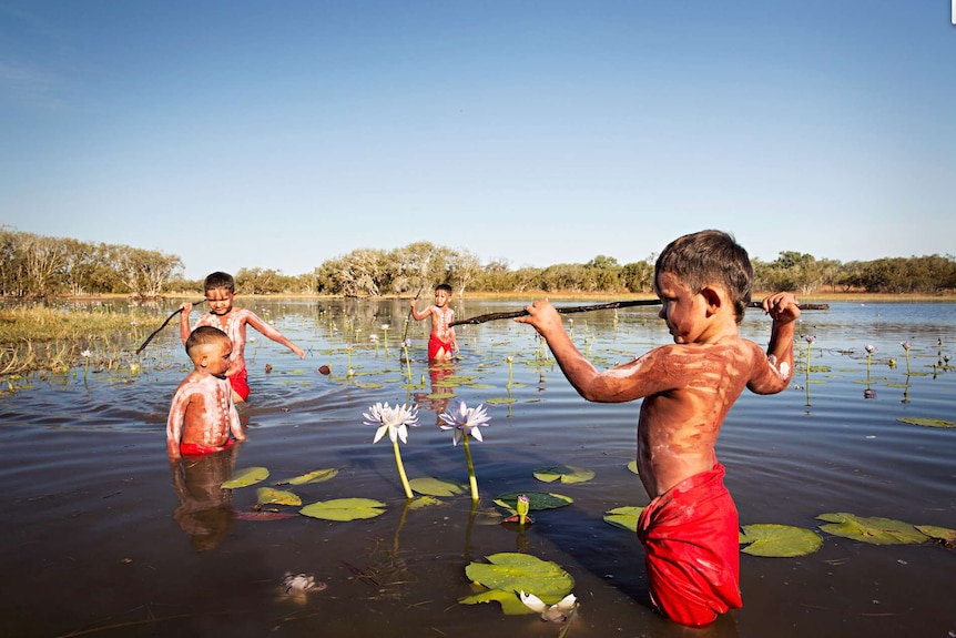 Indigenous children in a lake or creek with sticks imitating hunting.