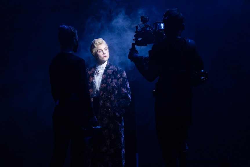 Blonde woman dressed as man in purple embroidered coat is onstage in plume of smoke surrounded by camera operators