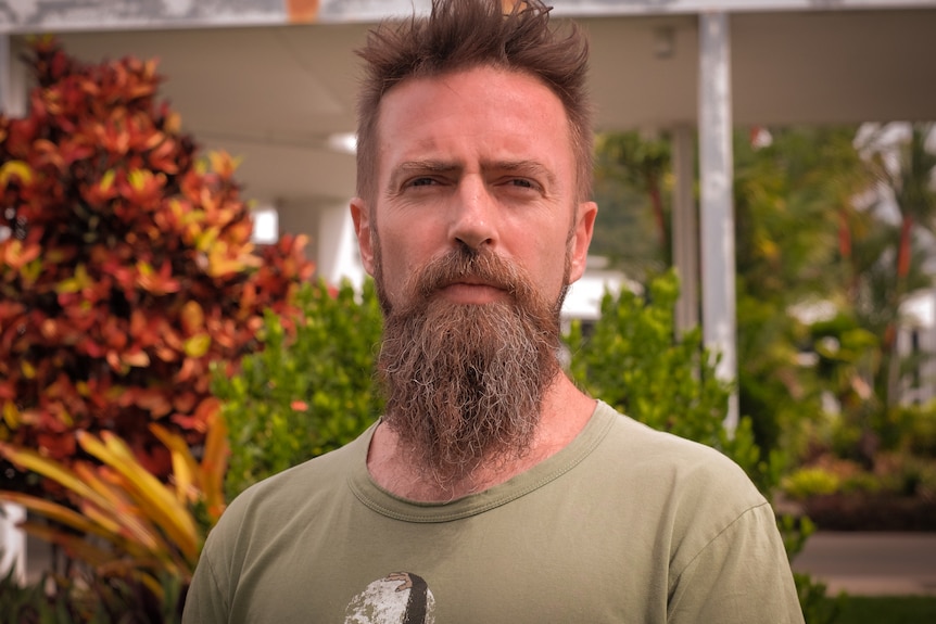A man with a beard and moustache stares at the camera while standing outside.