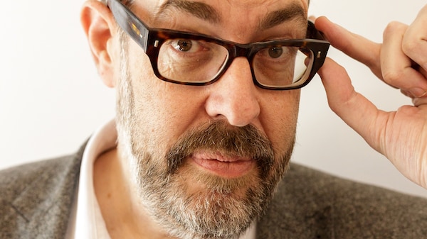 A middle-aged white man with a beard holds his fingers to his thick-rimmed glasses.