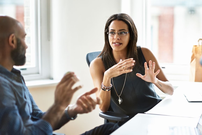 In an office environment, a woman gesticulates with her hands as she speaks with serious expression to a man who looks at her.