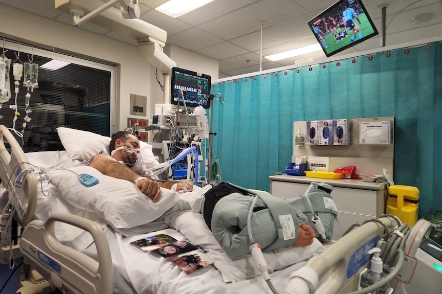 Wide shot of a man lying in a hospital bed with multiple wires and tubes, surrounding my machines and equipment