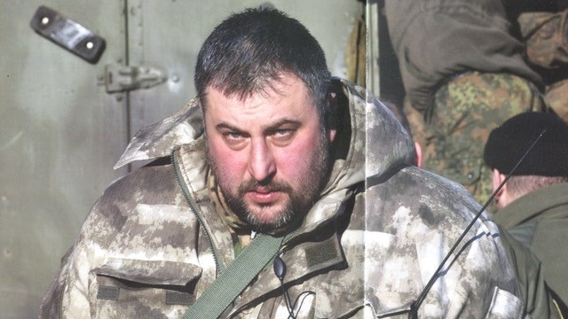 A heavyset man with close-cropped hair and a beard, dressed in military fatigues.