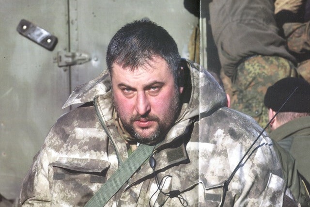 A heavyset man with close-cropped hair and a beard, dressed in military fatigues.