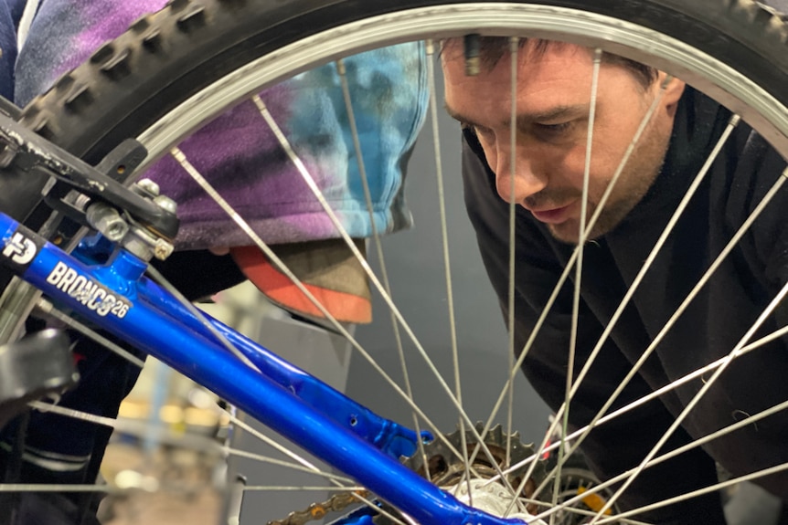 A man's face is half hidden behind the spokes of a bike wheel.