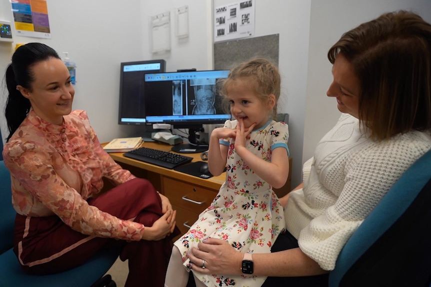 A woman speaks to a girl and another woman next to a computer