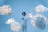 A woman stands in a blue room next to floating white clouds.