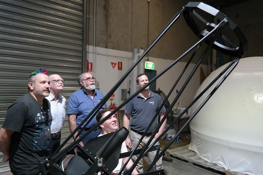 Five men, four standing and one sitting, look up happily at a large telescope.