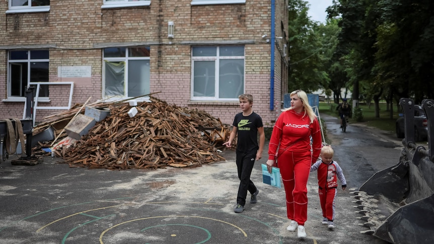 A woman, her teenage son and younger child in  the foreground of a damaged school building, and pile of broken planks