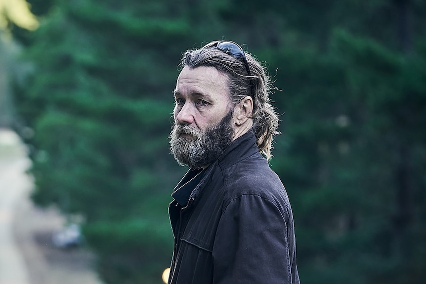 Middle-aged man white man with rustic beard, and long hair wearing black bomber jacket standing on a dirt road in bushy area.