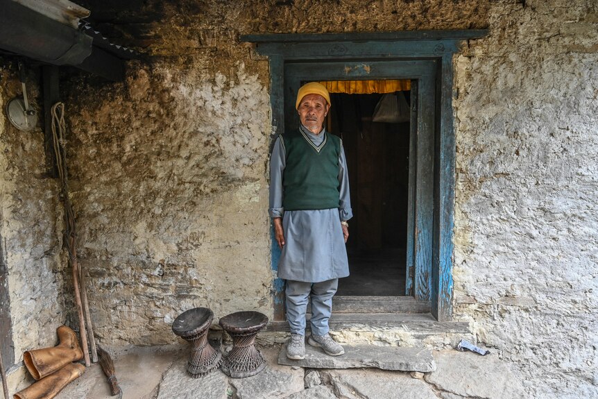 A man in a green vest with a yellow beanie stands in front of the wooden door into a mud brick building