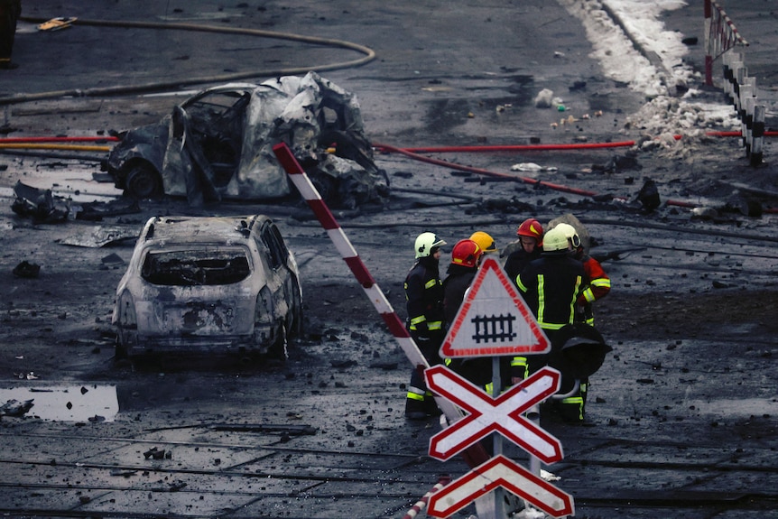 A group of fire service workers gather on a blackened, burnt street next to the wreckage of several cars.
