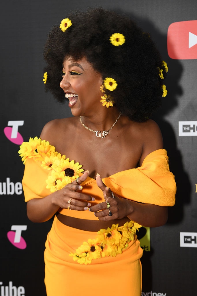 tsehay hawkins smiles while looking to the left of the shot wearing a yellow dress adorned with sunflowers and flower hairclips