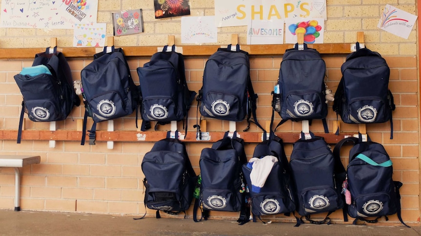 Schoolbags hanging on wall rack at St Hurmizd, with anniversary cards posted on wall above.