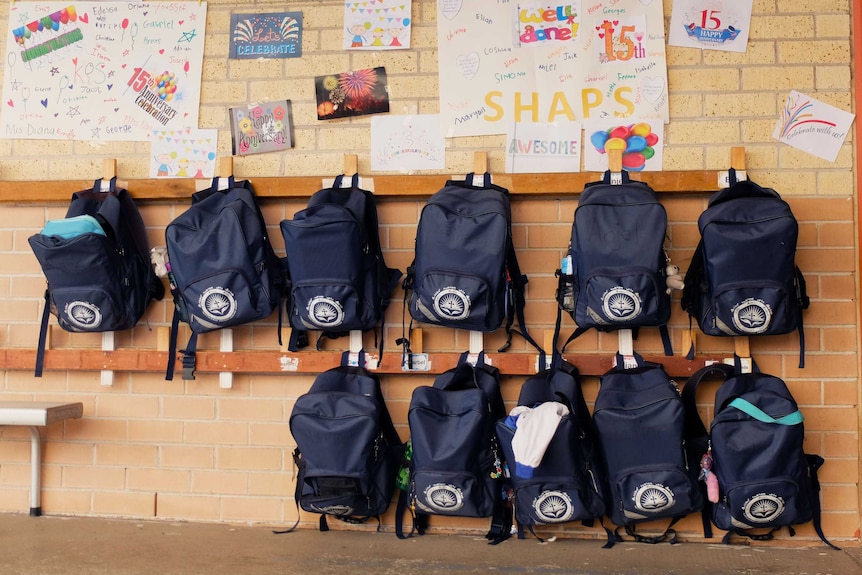 Schoolbags hanging on wall rack at St Hurmizd, with anniversary cards posted on wall above.