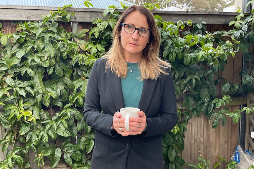 A woman with long brown hair and glasses holding a mug, standing in front of a vine