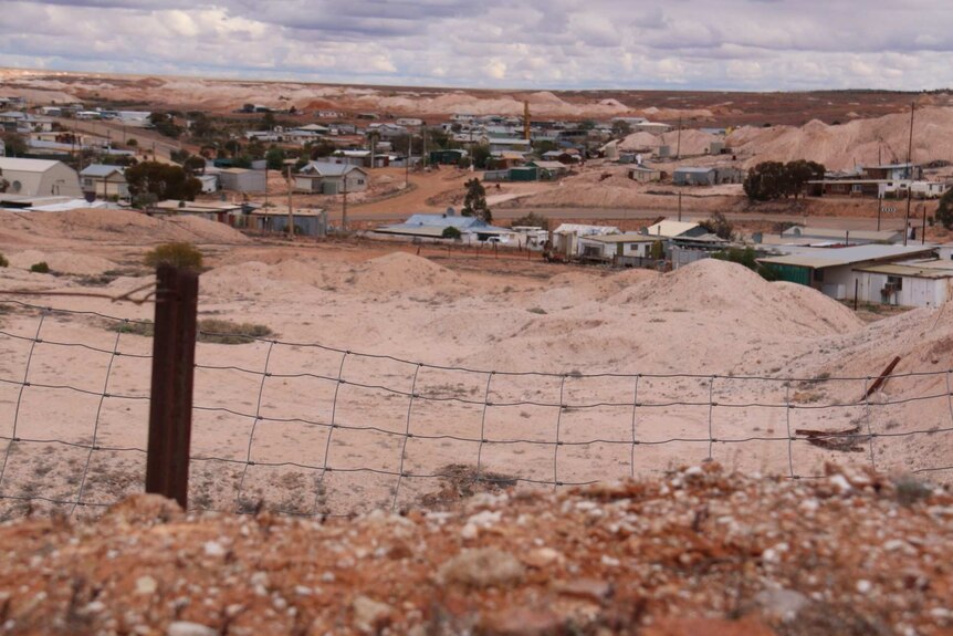 A wire fence surrounds a small historic town built on dark brown earth.