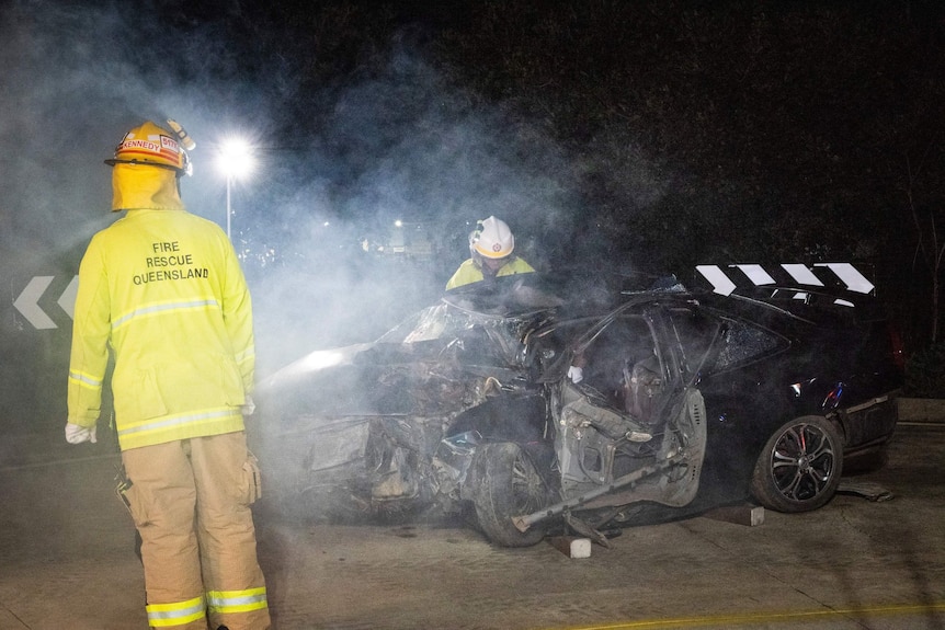 Two fire officers examining a small black sedan that has been wrecked in a crash.