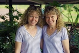 Identical twin sisters stand side-by-side smiling in identical outfits