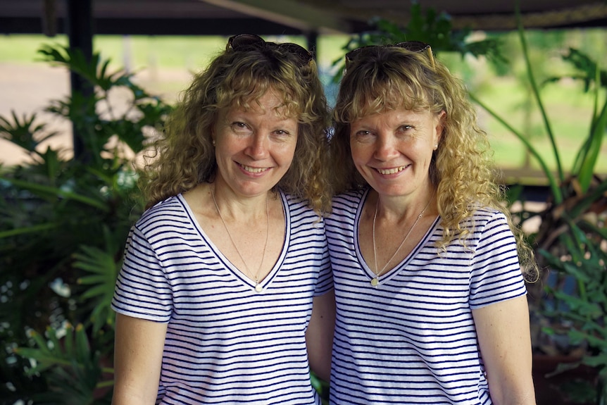 Identical twin sisters stand side-by-side smiling in identical outfits